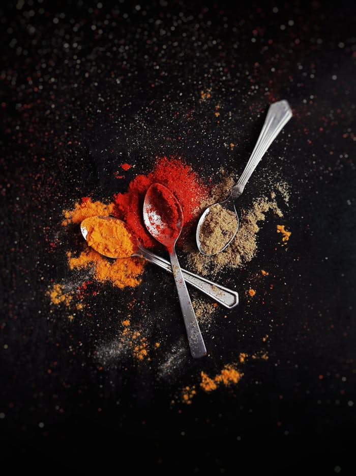 A creative flat lay of colorful spices - red, orange, and brown - with metal spoons on a dark background.